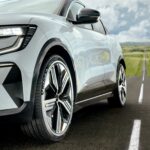 Coche de color blanco coronando una cuesta al final de una recta que atraviesa un campo verde